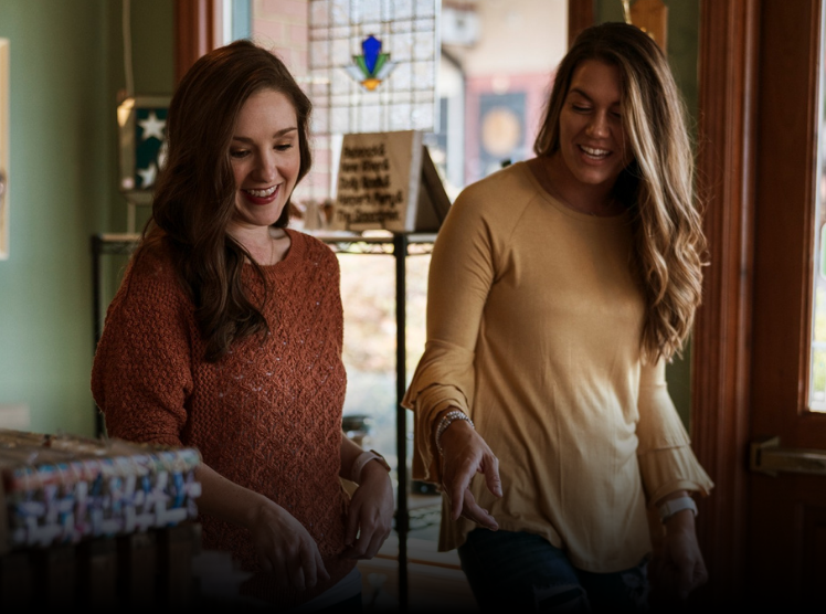Two smiling women standing inside a shop, looking down at something out of frame.