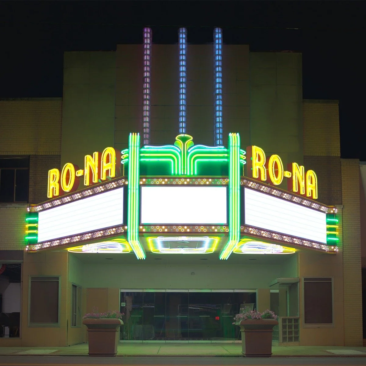 Neon-lit theater marquee for 'Roa Na' with empty white marquee space in the center, flanked by potted plants, at night.