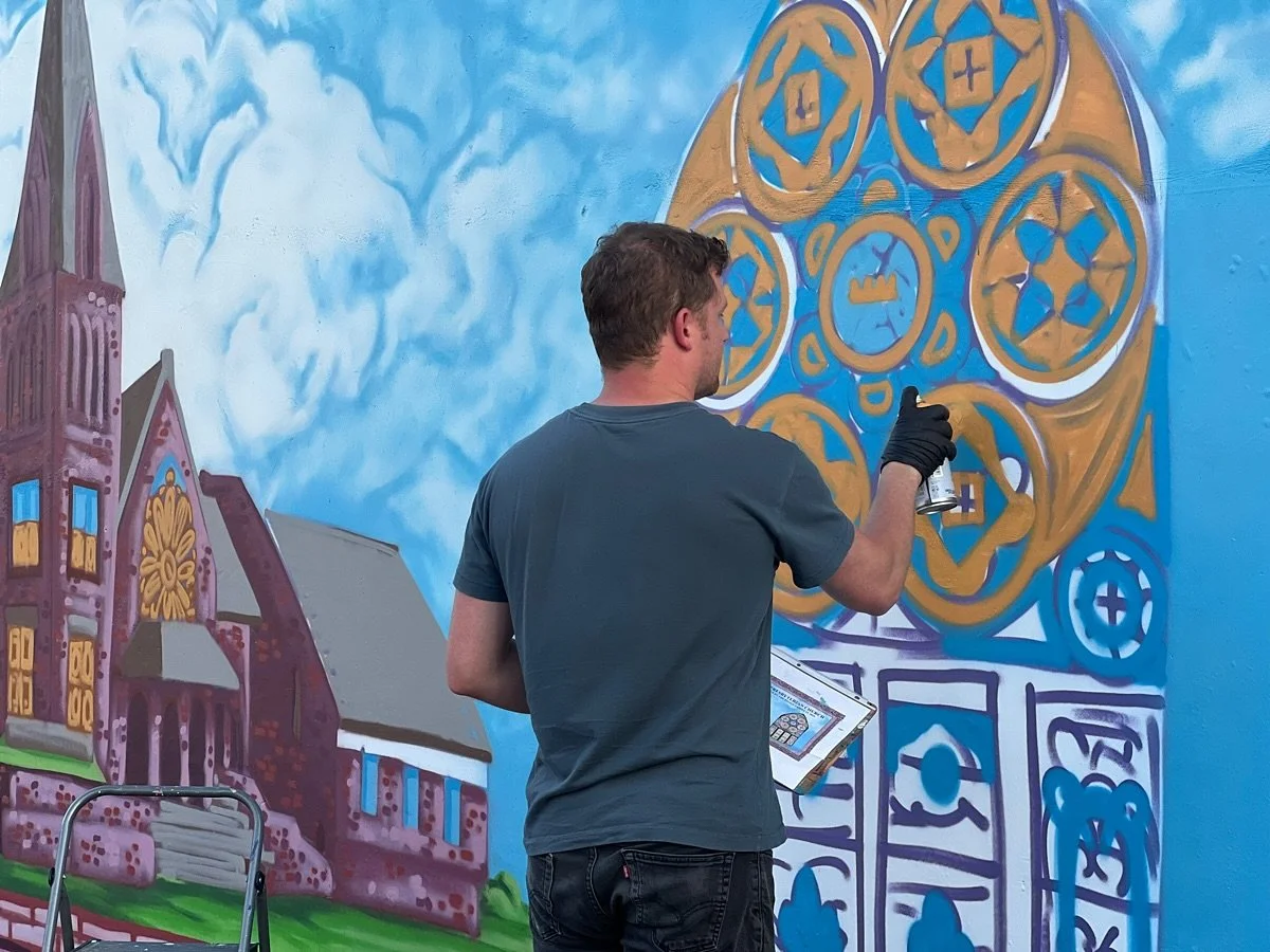A man with dark hair, wearing a blue shirt and black gloves, is spray painting a colorful mural on a blue wall. The mural features a large geometric design with symbols and a church with a tower, windows, and a door in front of a sky with clouds.