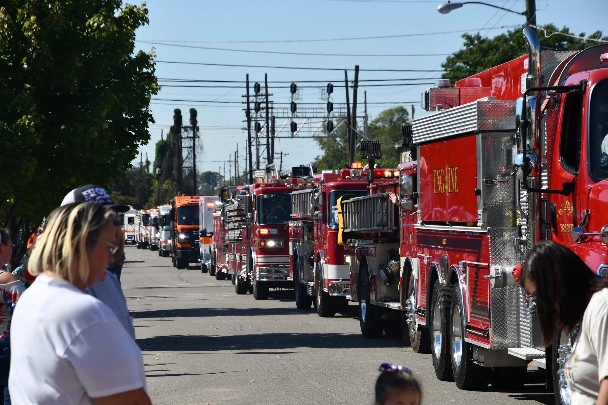Line of red fire trucks parked on the street with onlookers observing, power lines and trees in the background.