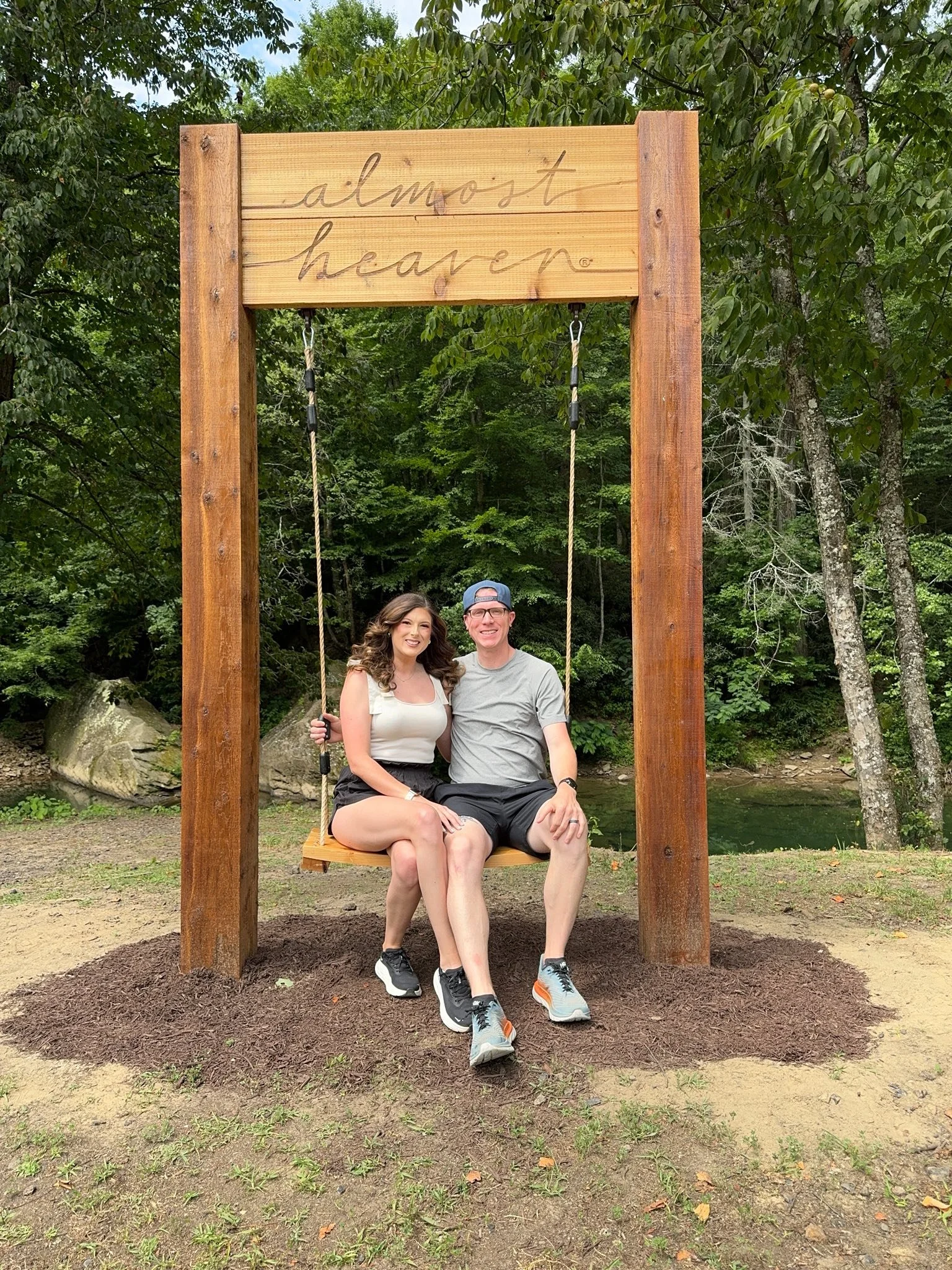 A smiling couple sitting on a large wooden swing in a lush green park, with trees and a body of water in the background. The swing has a sign that reads "almost heaven."