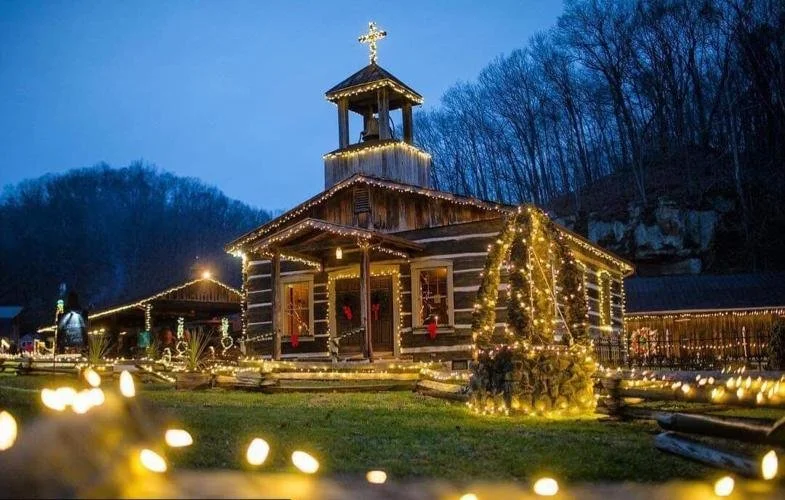 A small wooden church decorated with Christmas lights and ornaments, set outdoors during dusk, surrounded by festive lights and trees.