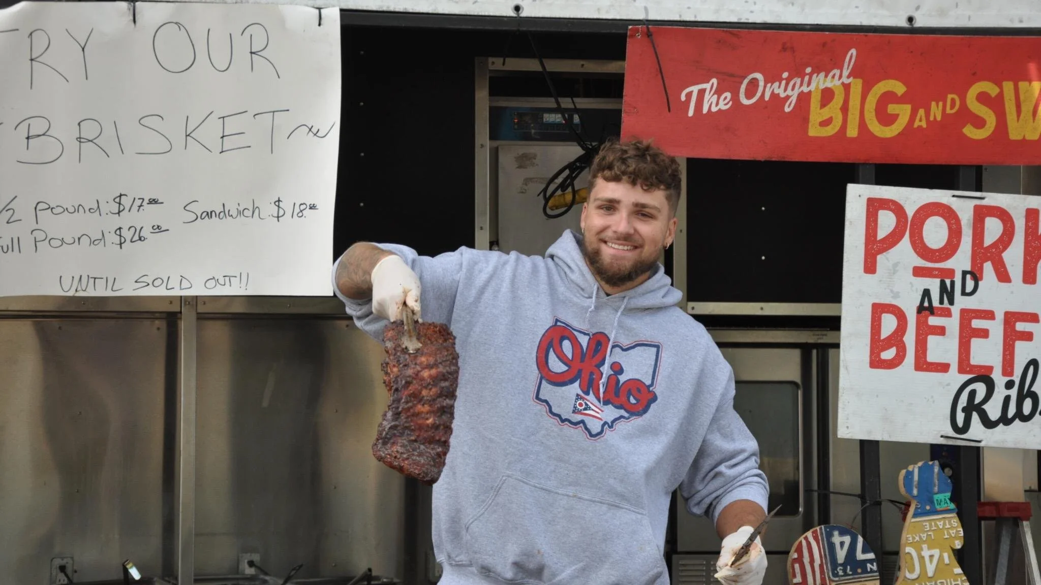 A young man smiling and holding a large barbecue pork rib at a food stand. Behind him are signs advertising brisket and pork rib sandwiches.