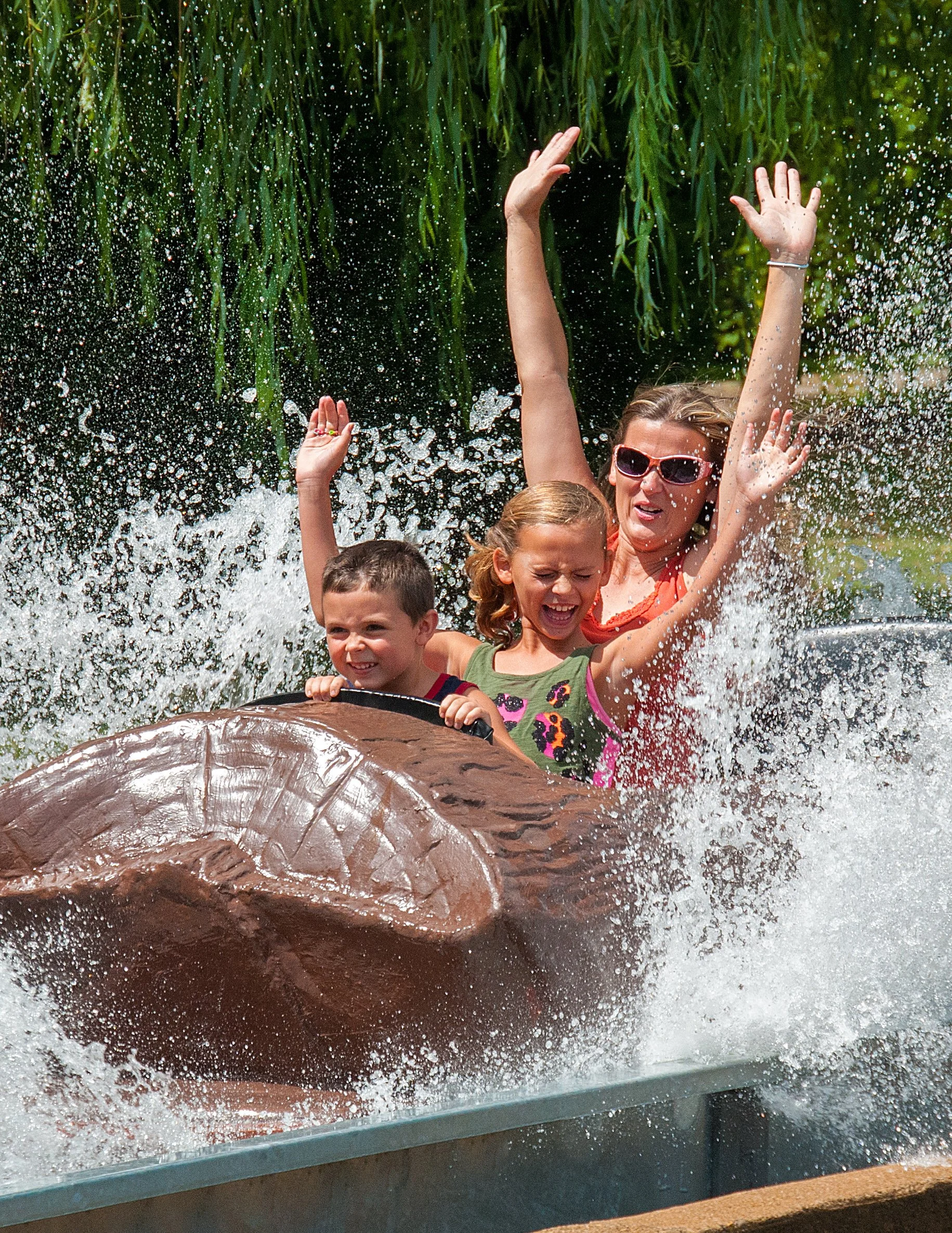 A woman and two children riding a log flume at an amusement park, splashing water and smiling.