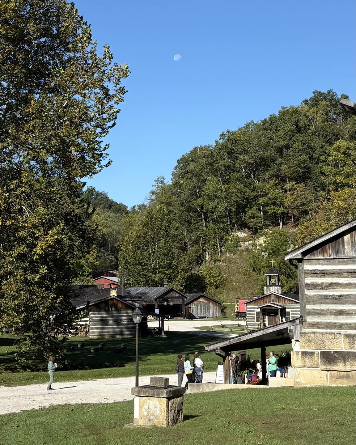 A historic village with wooden buildings and a group of people gathering outside, surrounded by trees and a clear blue sky with a visible moon.