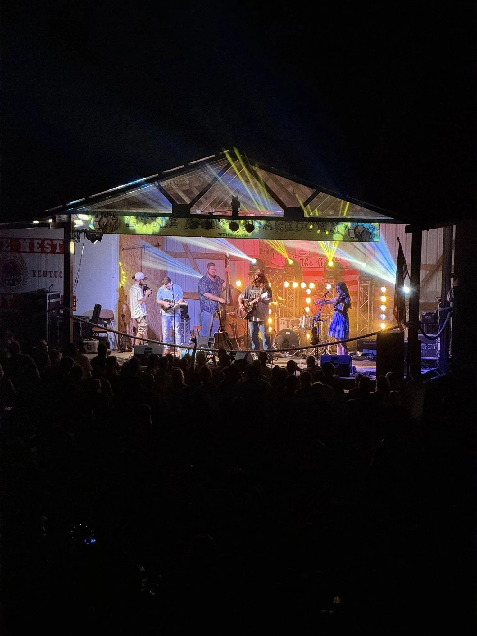 A live music concert on a stage at night with a band playing instruments, colorful stage lights, and an audience watching.