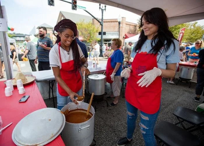 Two women wearing red aprons and gloves serving chili at an outdoor food festival. One woman is scooping chili into a bowl, and the other is smiling, standing next to a large pot.