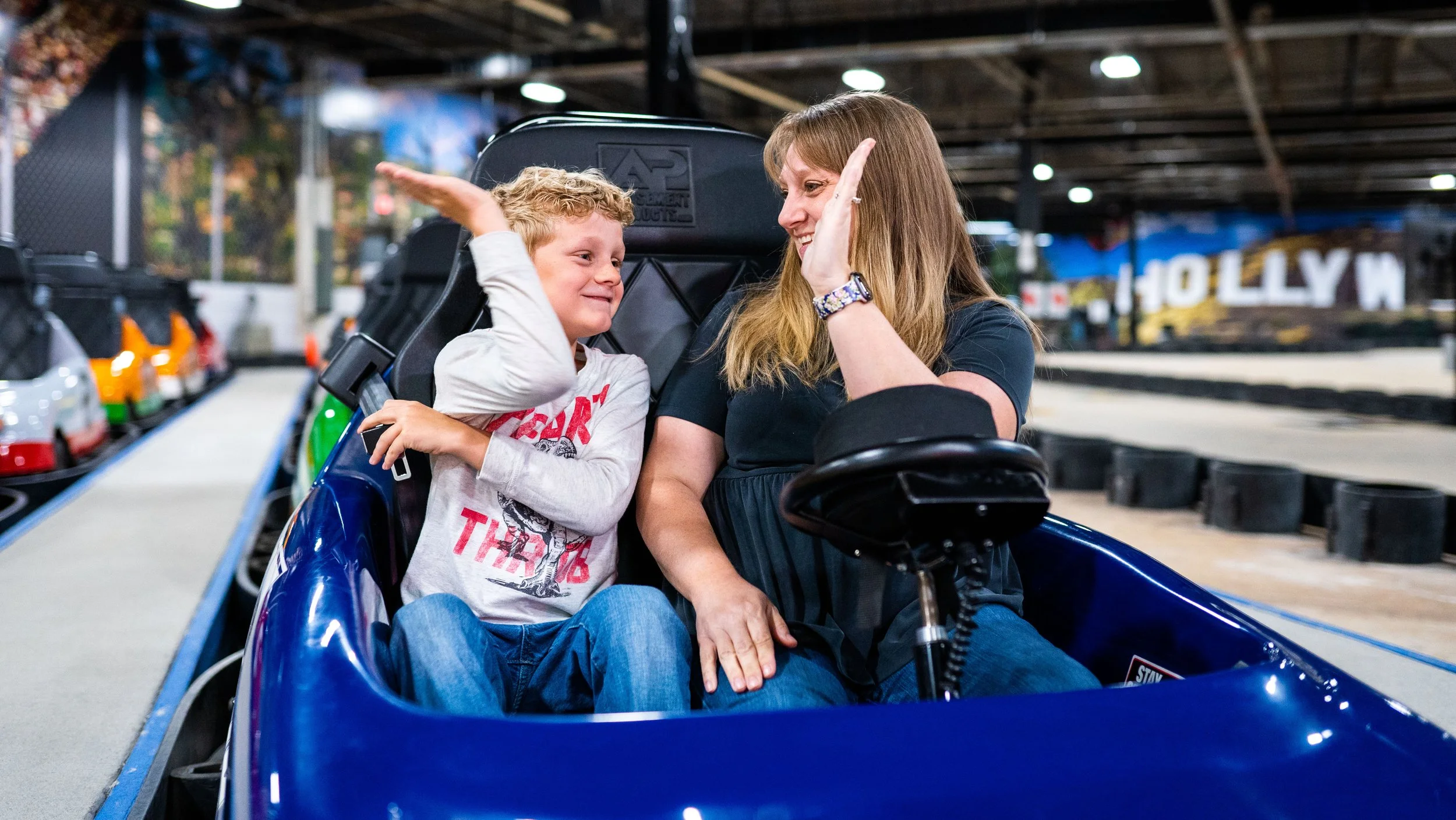 A woman and a young boy sitting in a blue go-kart, giving each other high fives and smiling at each other in an indoor go-kart racing track.