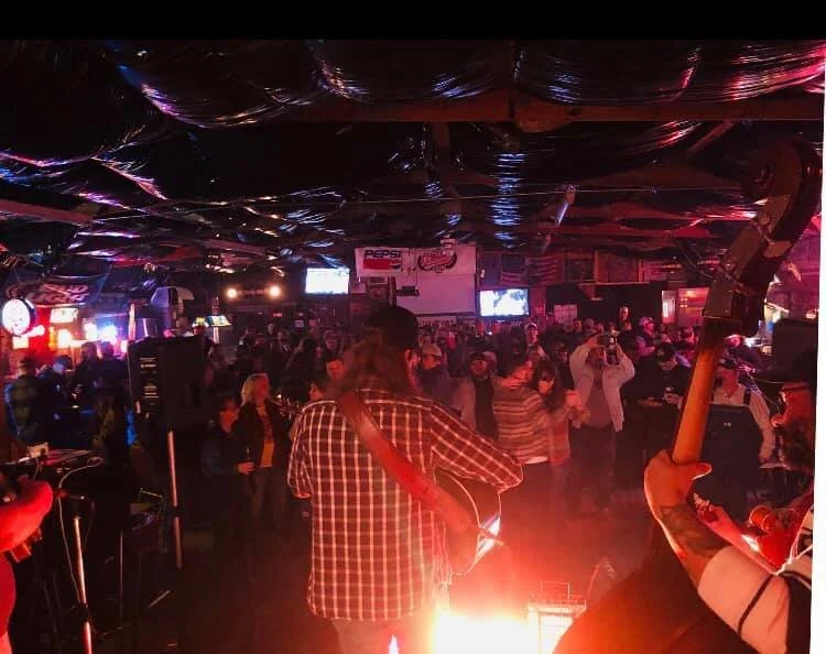 Crowd of people at a lively indoor concert or event with stage lights and musical instruments.