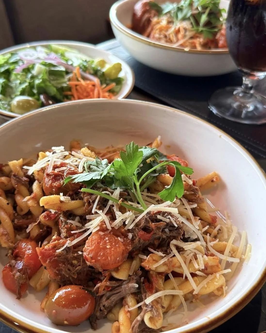 A plate of baked pasta with tomatoes, herbs, and grated cheese, garnished with fresh parsley, with a side salad and a glass of red wine in the background.