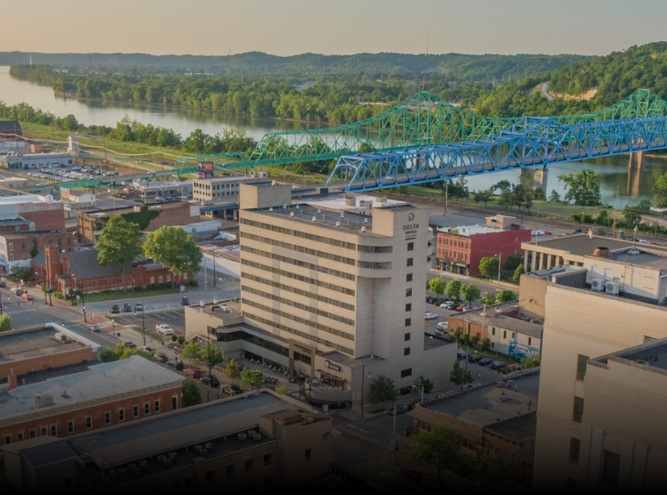 An aerial view of a city with a river, a bridge painted in green and blue, and various buildings including a tall hotel with the Delta logo, surrounded by trees and roads.