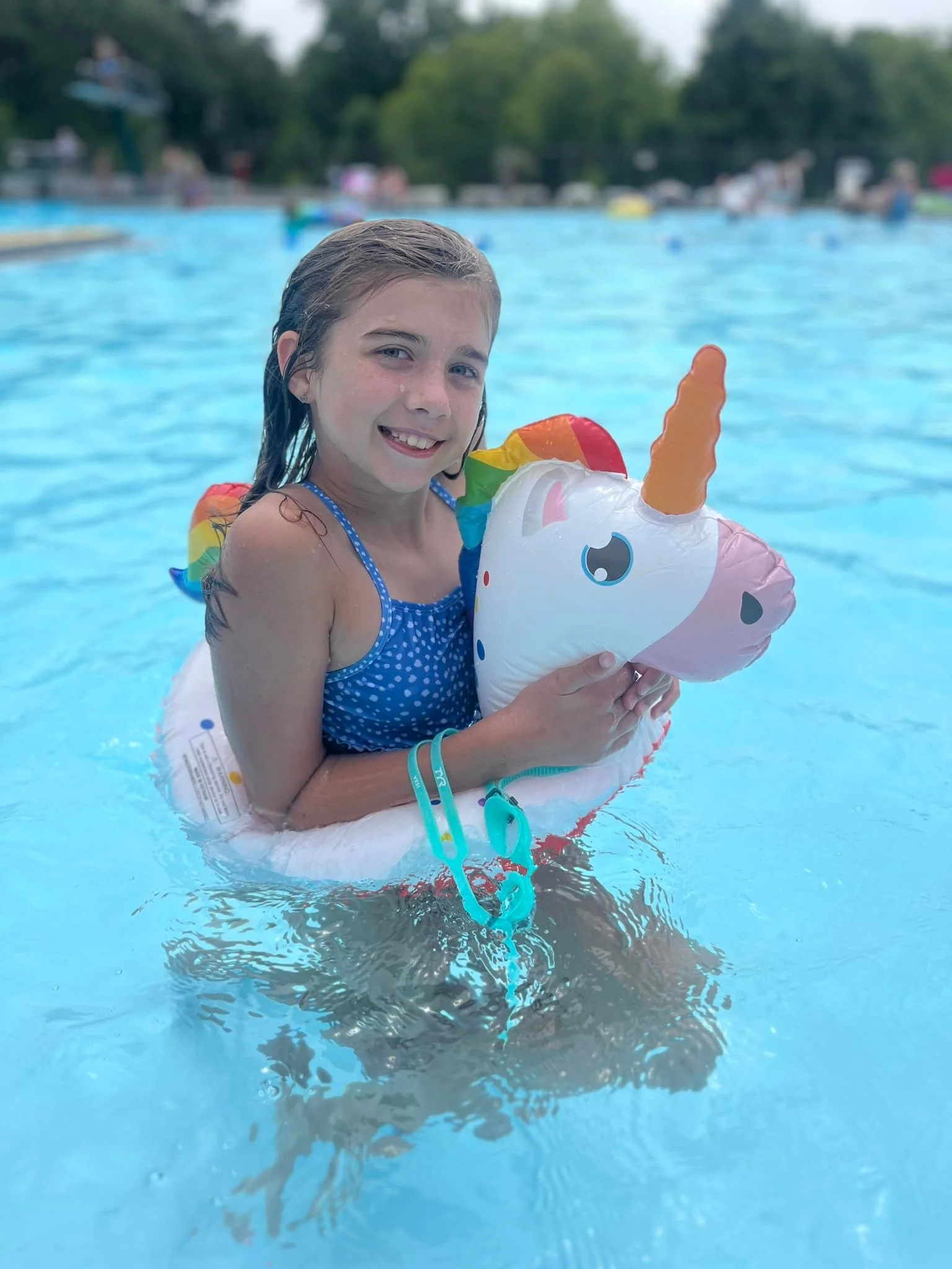 Young girl in a blue polka-dot swimsuit smiling in a swimming pool, holding a unicorn-shaped inflatable float.