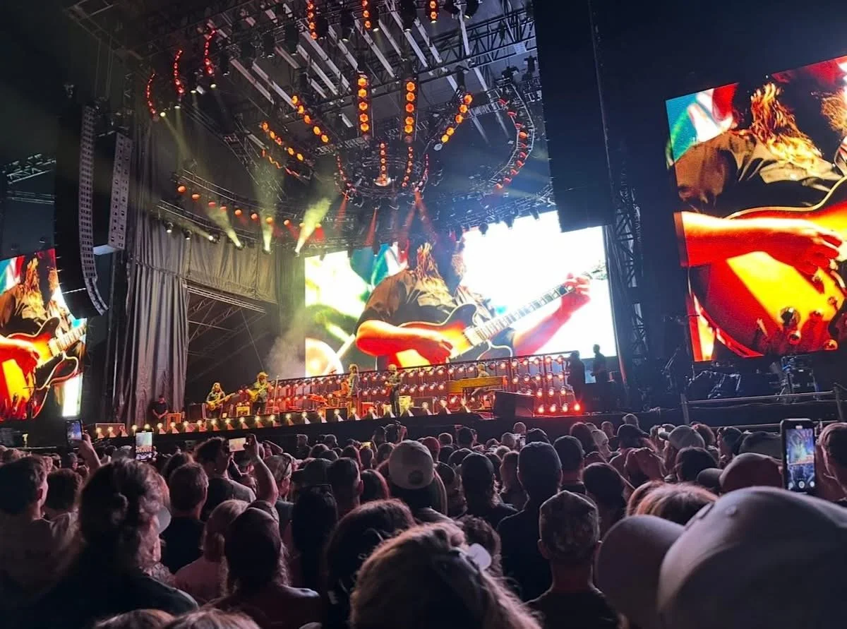 A live concert with a large audience, colorful stage lighting, and big screens showing a guitarist playing an electric guitar.