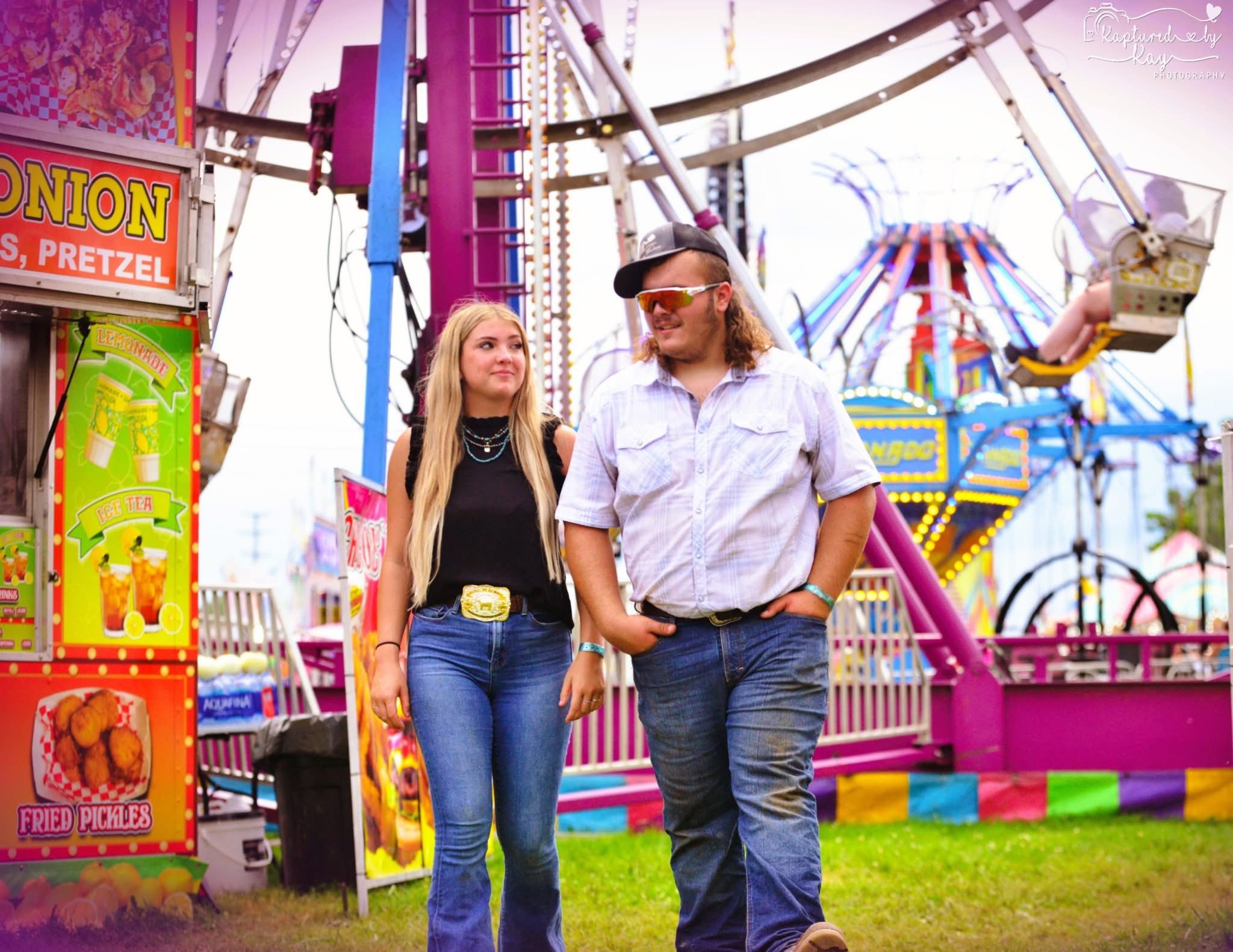 A young woman and man walking at a fairground with colorful amusement rides in the background, including a ferris wheel. The woman has long blonde hair, wears a black top, blue jeans, a decorative belt, and necklaces. The man has long hair, wears a white shirt, blue jeans, sunglasses, a cap, and has his hand in his pocket.