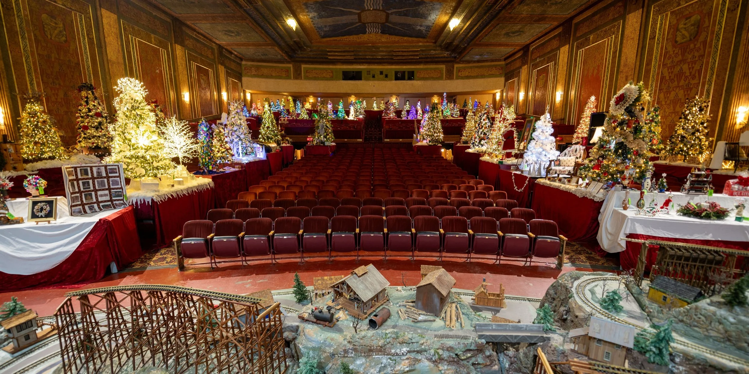 An ornately decorated theater with numerous Christmas trees, lights, and holiday decorations. There is a model train set and miniature buildings on the floor in the foreground.