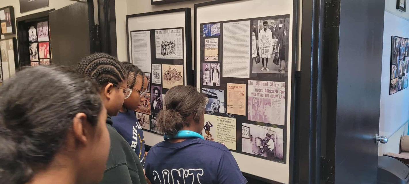 Four women standing in front of a historical display at a museum, reading framed informational posters and photographs.