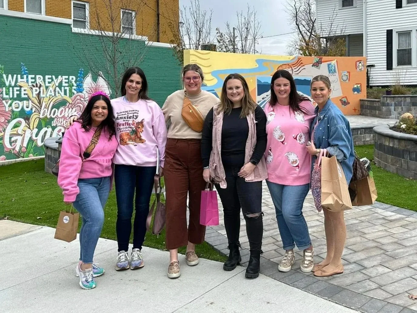Group of six women standing outdoors in front of colorful mural, holding shopping bags, smiling at the camera.