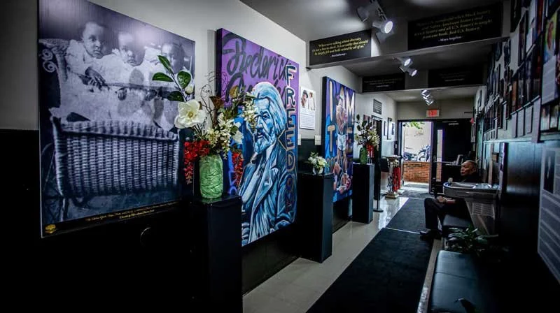Art gallery interior with portraits on the wall, floral displays on pedestals, and a person sitting near a piano.