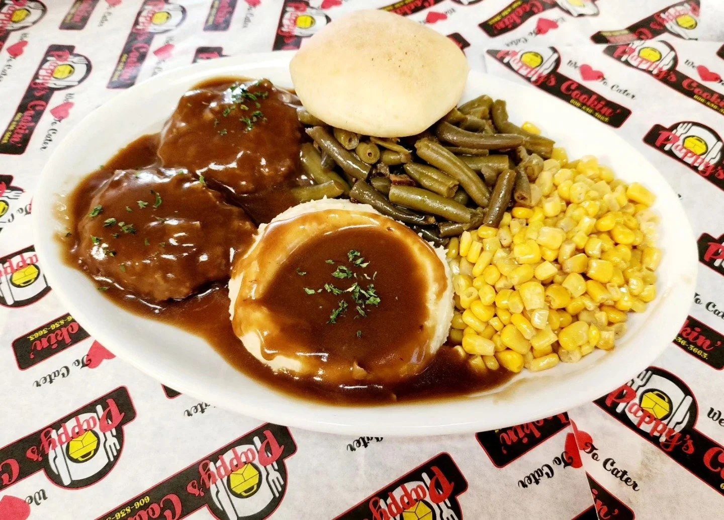 Plate of breaded chicken covered in gravy, mashed potatoes with gravy, green beans, and corn, with a bread roll on the side, on a table with restaurant branded paper.