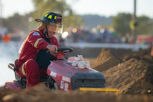 A firefighter wearing a red uniform and helmet, sitting on a small race car, holding a teddy bear, on a dirt track with a crowd in the background.