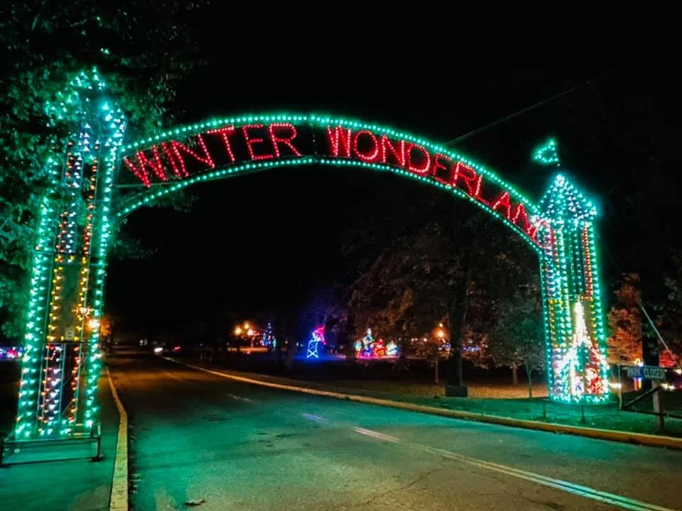 An illuminated archway with the words 'WINTER WONDERLAND' in red lights, decorated with colorful Christmas lights, at night.