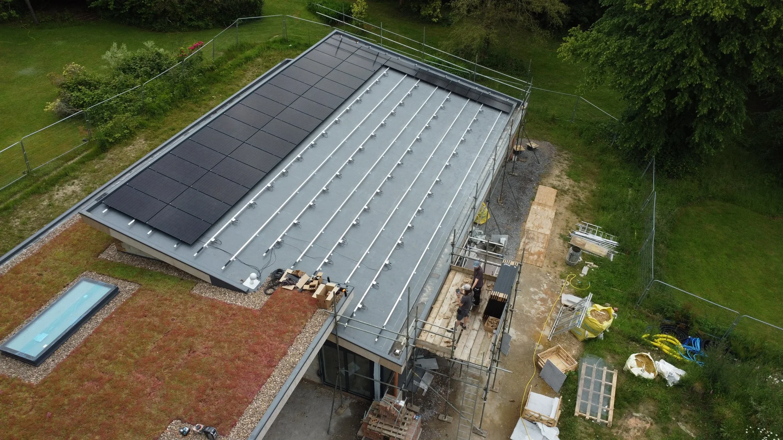 Aerial view of a modern house under construction with solar panels on the roof, scaffolding around the building, and construction materials scattered around the yard.