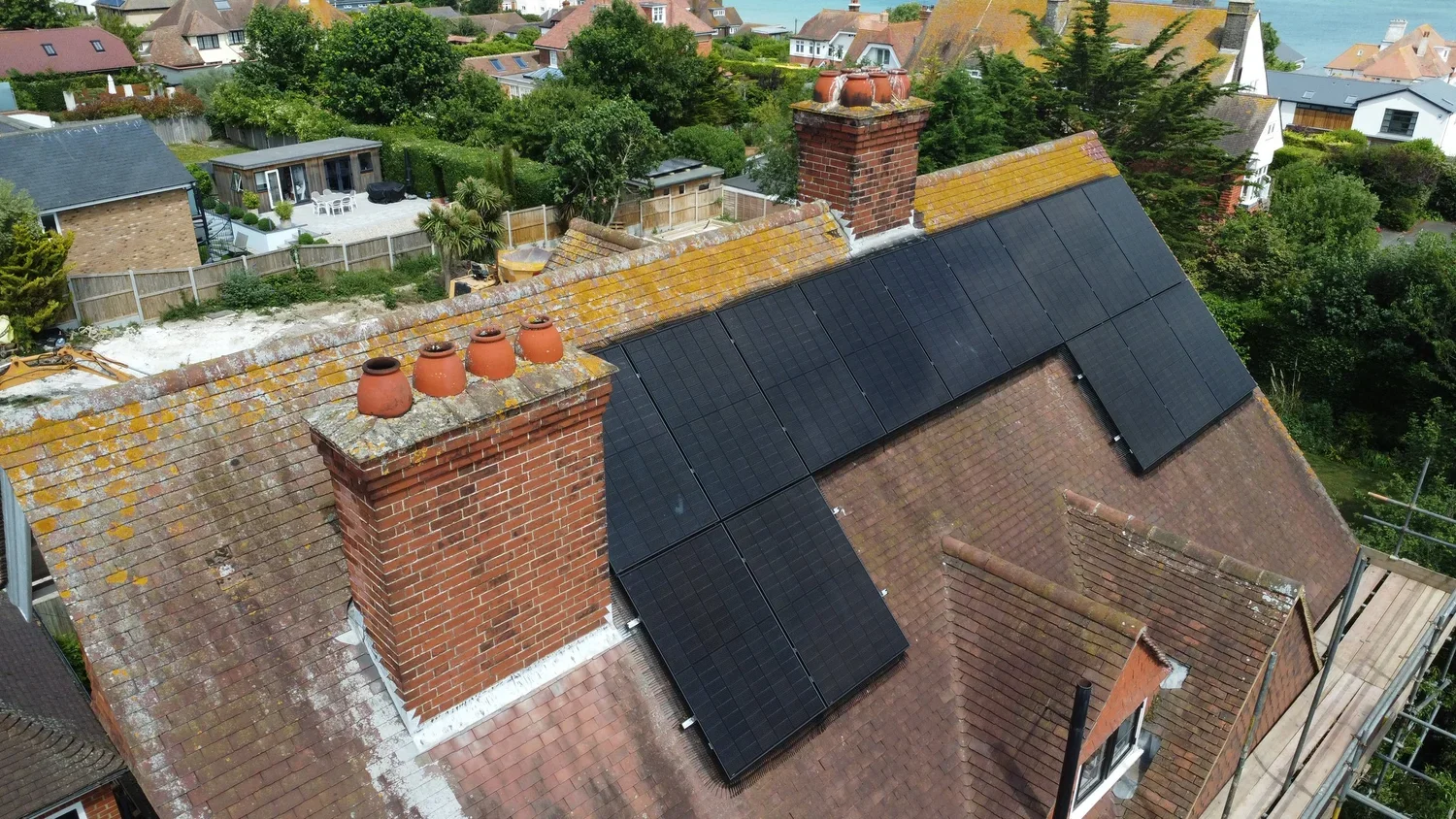 Aerial view of a house with black solar panels installed on the roof, brick chimneys, and an aged tile roof with moss. The background shows neighboring houses, trees, and a blue sky.