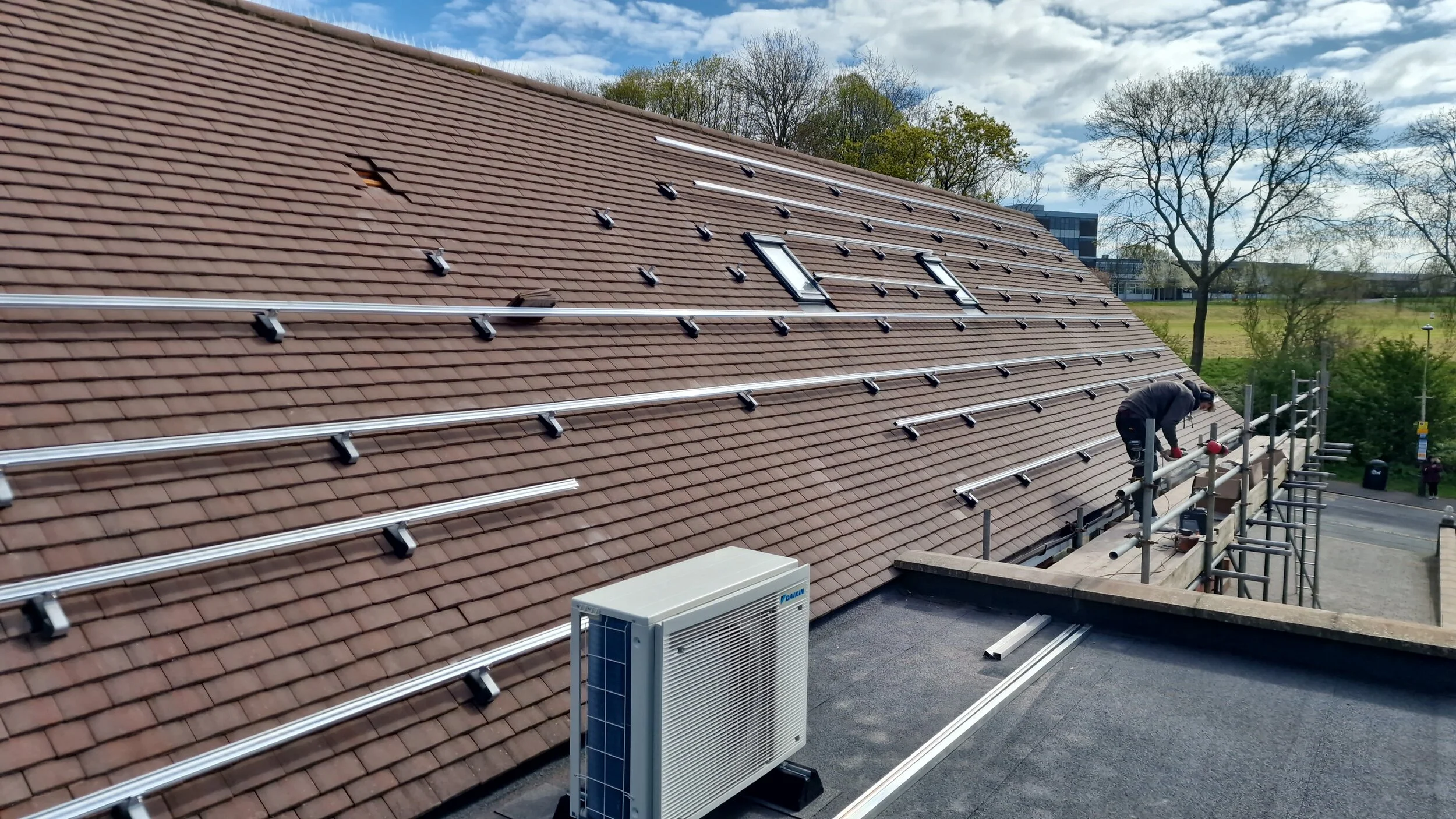 A worker installing roof solar mounting rails on a tiled roof with skylights, with scaffolding and trees in the background.