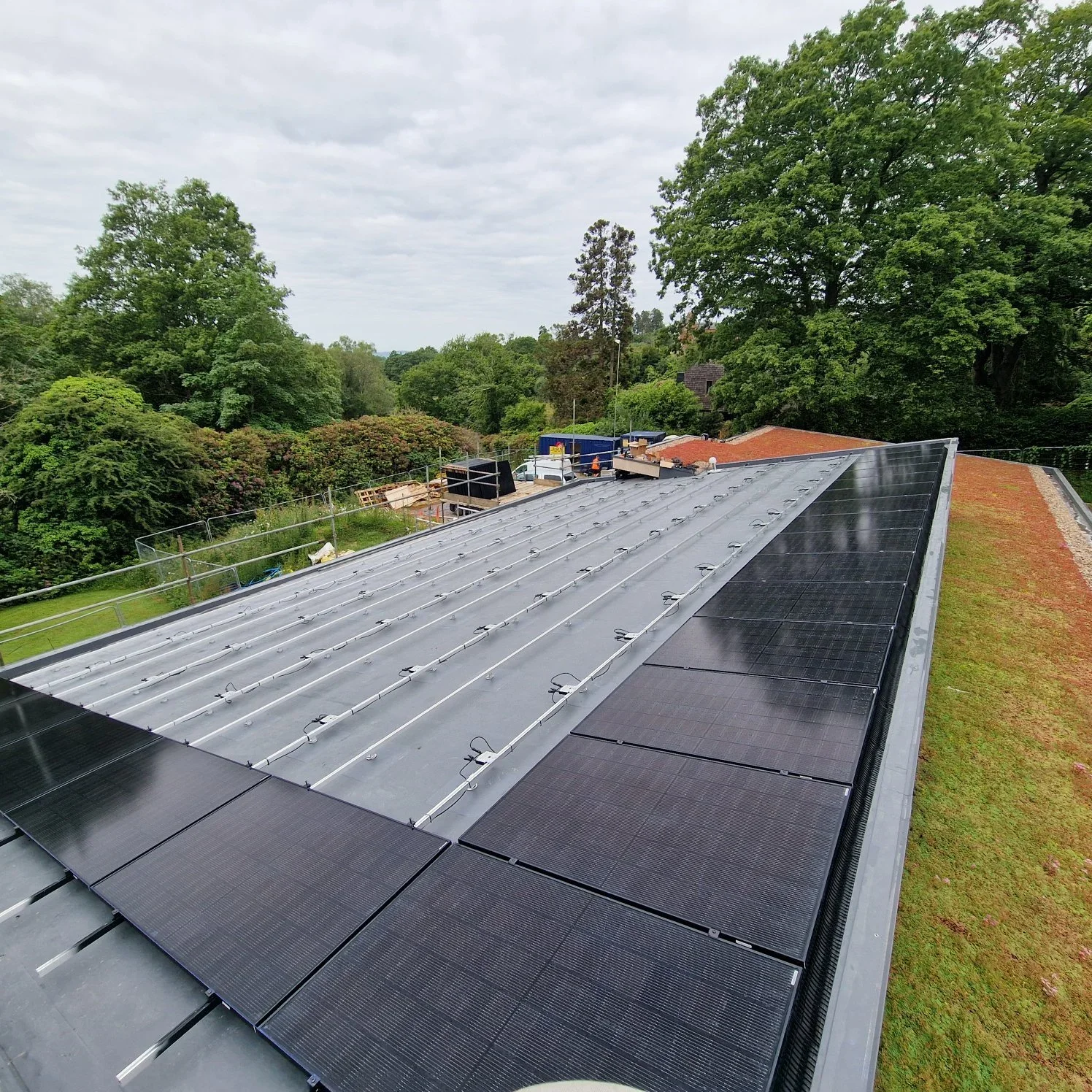 Rooftop with solar panels and metal roofing, surrounded by trees and overcast sky.