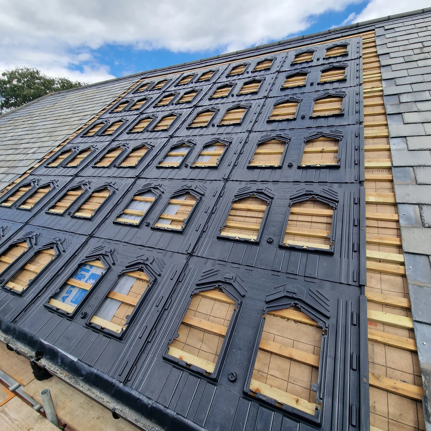 Roof under construction with black underlayment and wooden framing, additional slate shingles to the right, green trees in the background, and a partly cloudy blue sky.