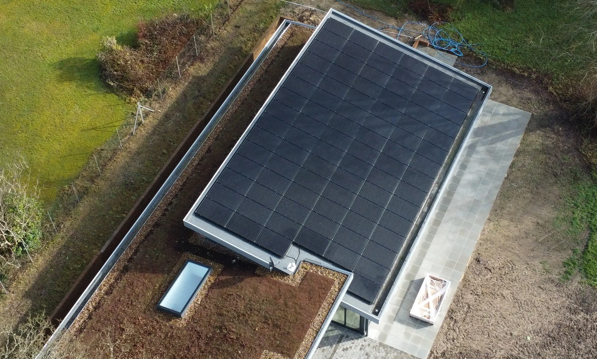 An aerial view of a modern house with a roof equipped with solar panels, surrounded by a green lawn, trees, and a paved patio area.