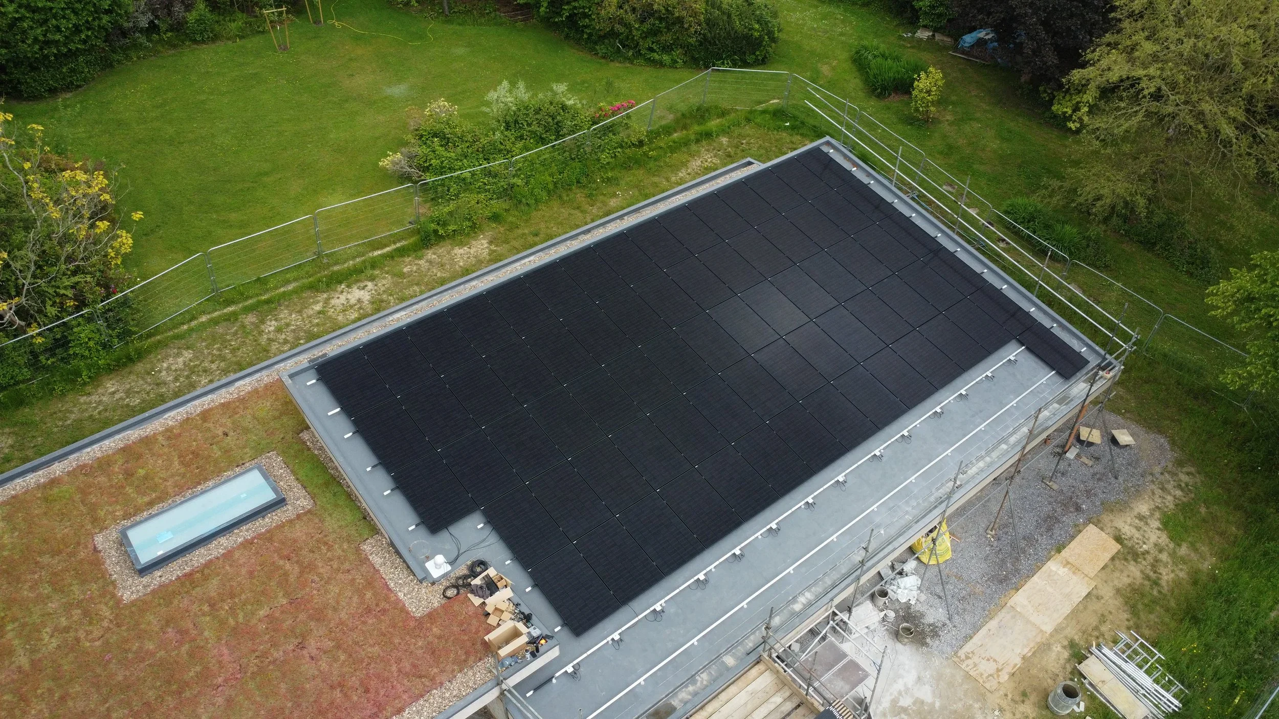 A rooftop with black solar panels installed, surrounded by a metal safety railing, with a backyard that includes a small rectangular pool, green grass, and trees.