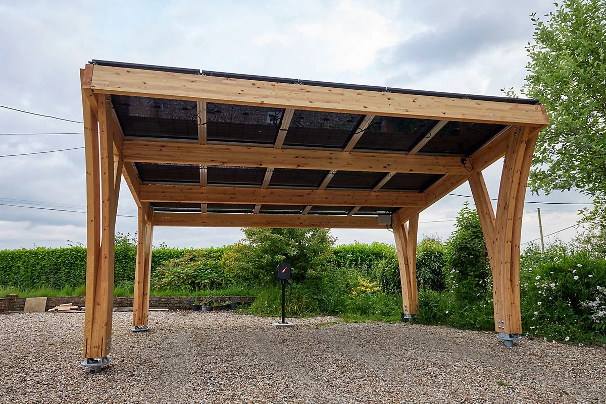 Wooden carport with solar panels installed on the roof, situated on gravel ground with green bushes and a tree in the background under an overcast sky.