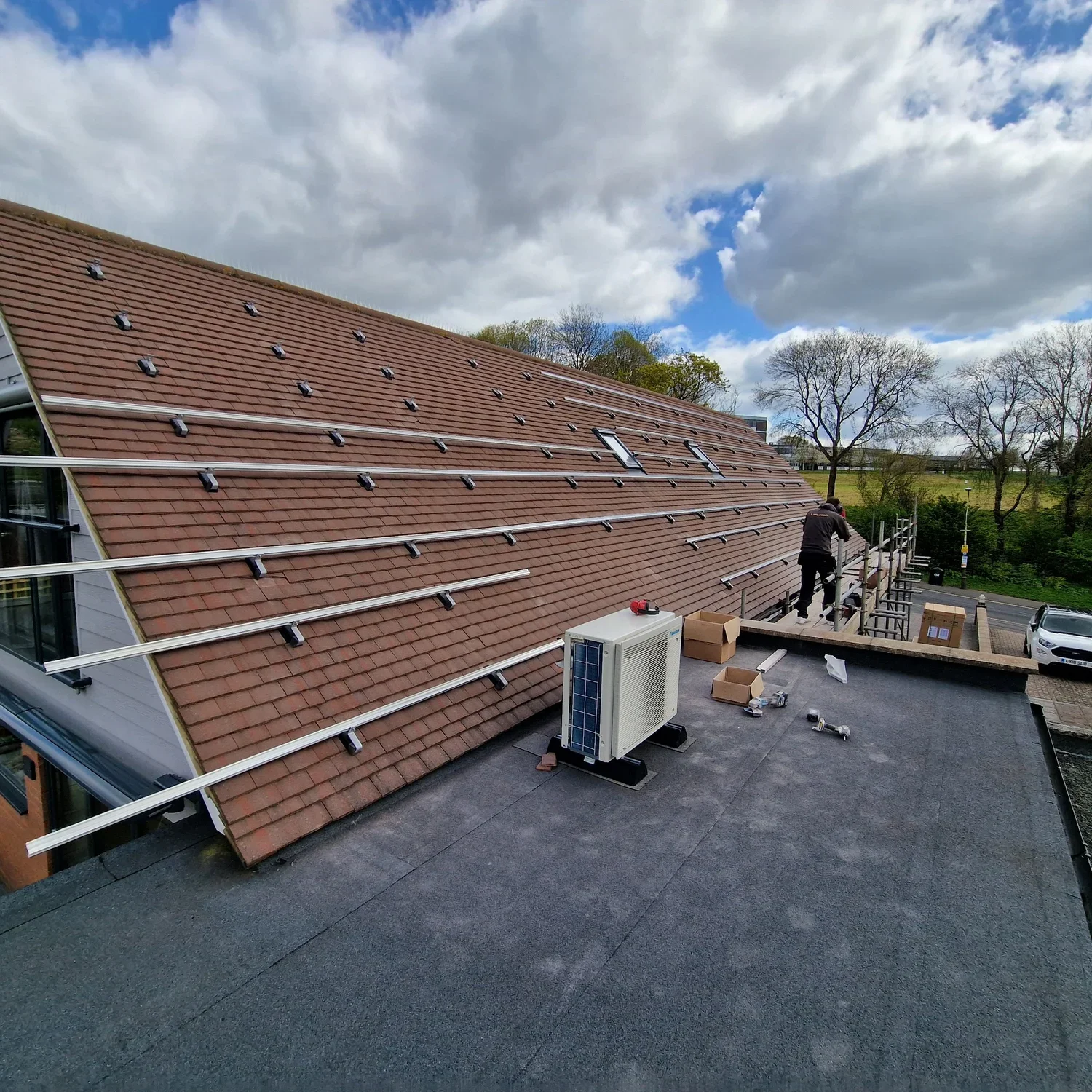 Person working on scaffolding installation on a sloped roof with solar panel mounting rails, under cloudy sky, in a residential area with trees and parked cars.