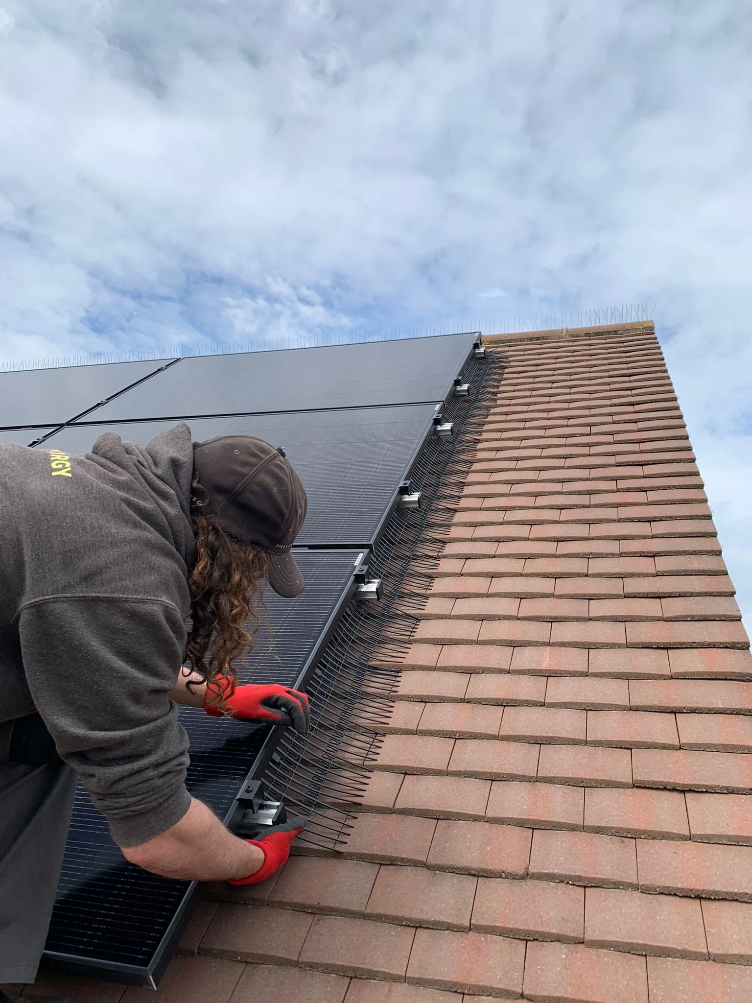 A worker installing solar panels on a brick roof, wearing gloves and a cap, with a cloudy sky in the background.