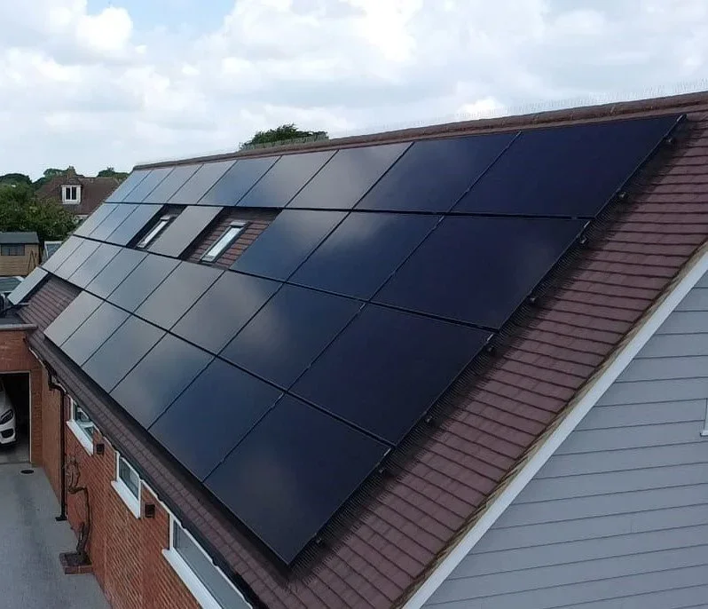 House roof covered with black solar panels, with a neighboring house and cloudy sky in the background.