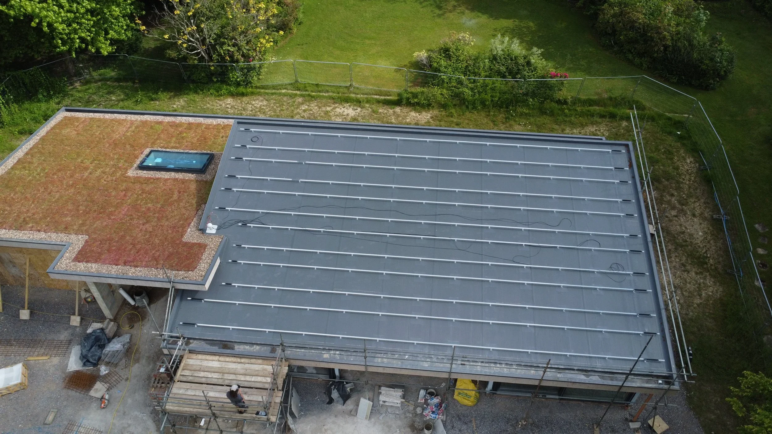 Aerial view of a building under construction with a metal roof, a smaller section with a green roofing material, and a skylight window. The building is surrounded by grass and trees.