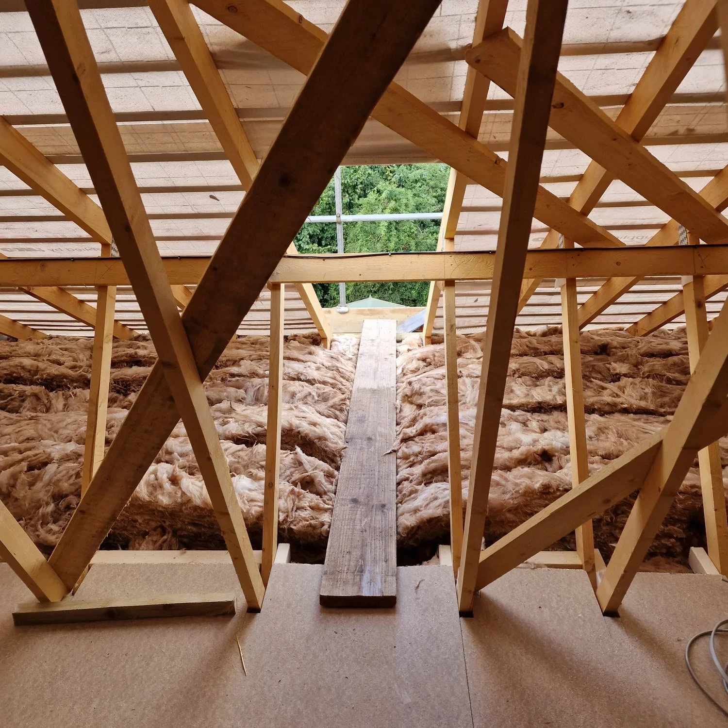 View of a house under construction with exposed wooden framing and insulation, and an opening at the top showing green trees outside.