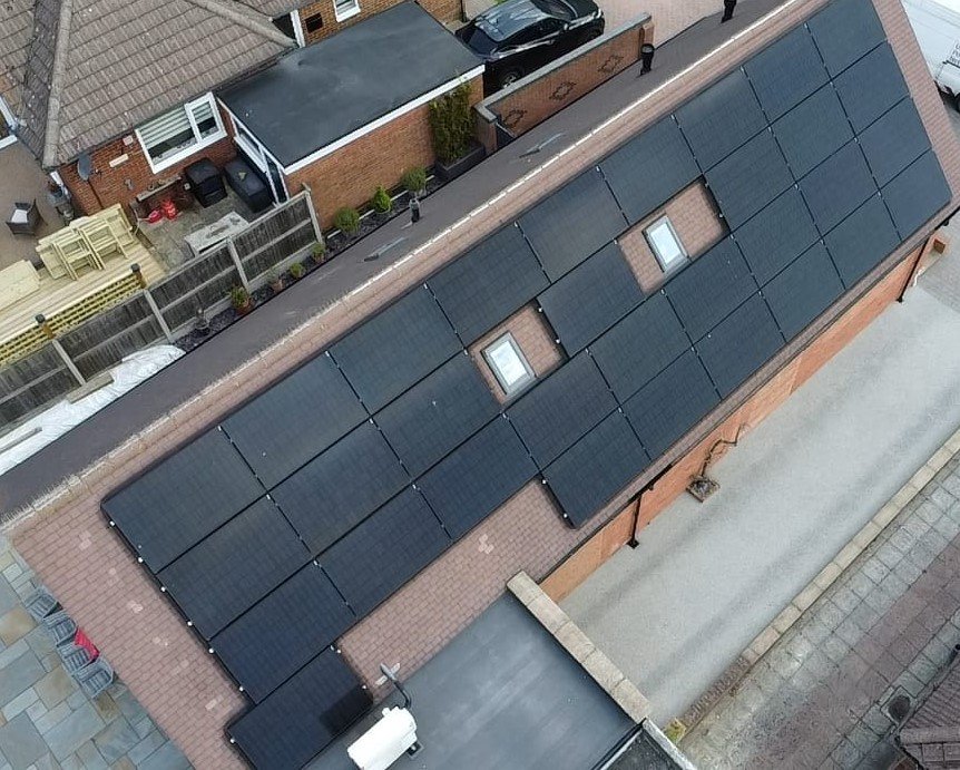An aerial view of a building rooftop covered with solar panels, with neighboring houses and a small parking area visible.