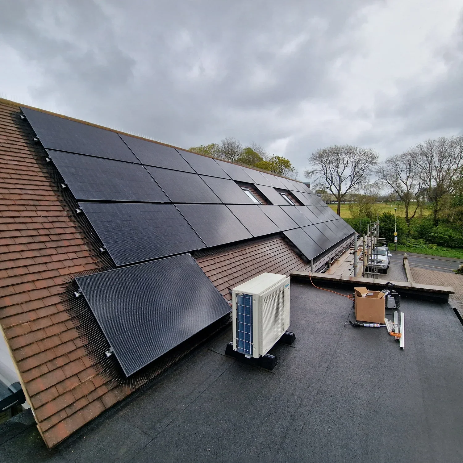 Solar panels installed on a sloped roof, with an air conditioning unit and tools nearby on a flat roof. Trees and a parking lot are visible in the background.