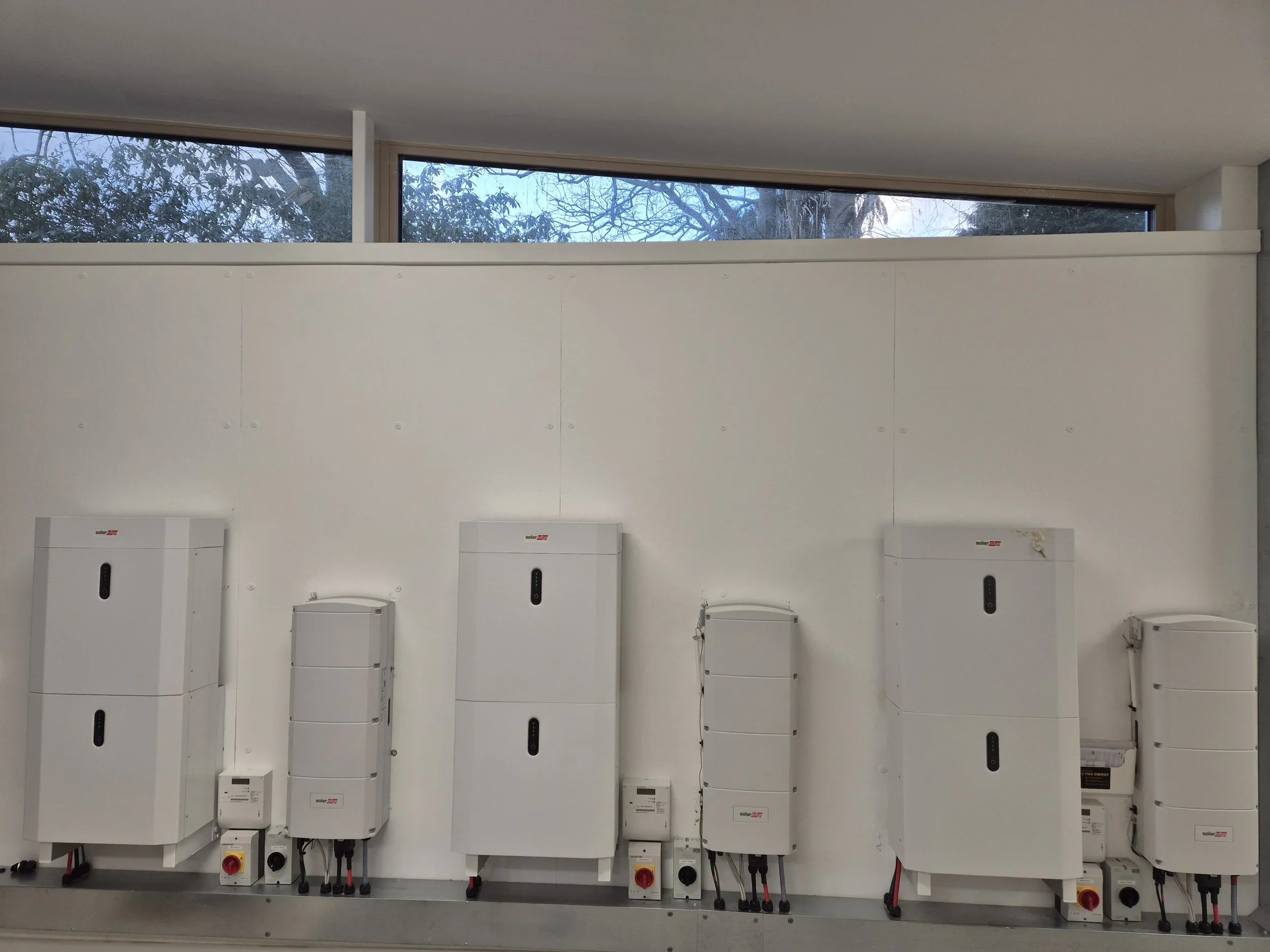 Installation of multiple solar inverters on a white wall, with visible wiring and electrical components, and windows showing trees outside.