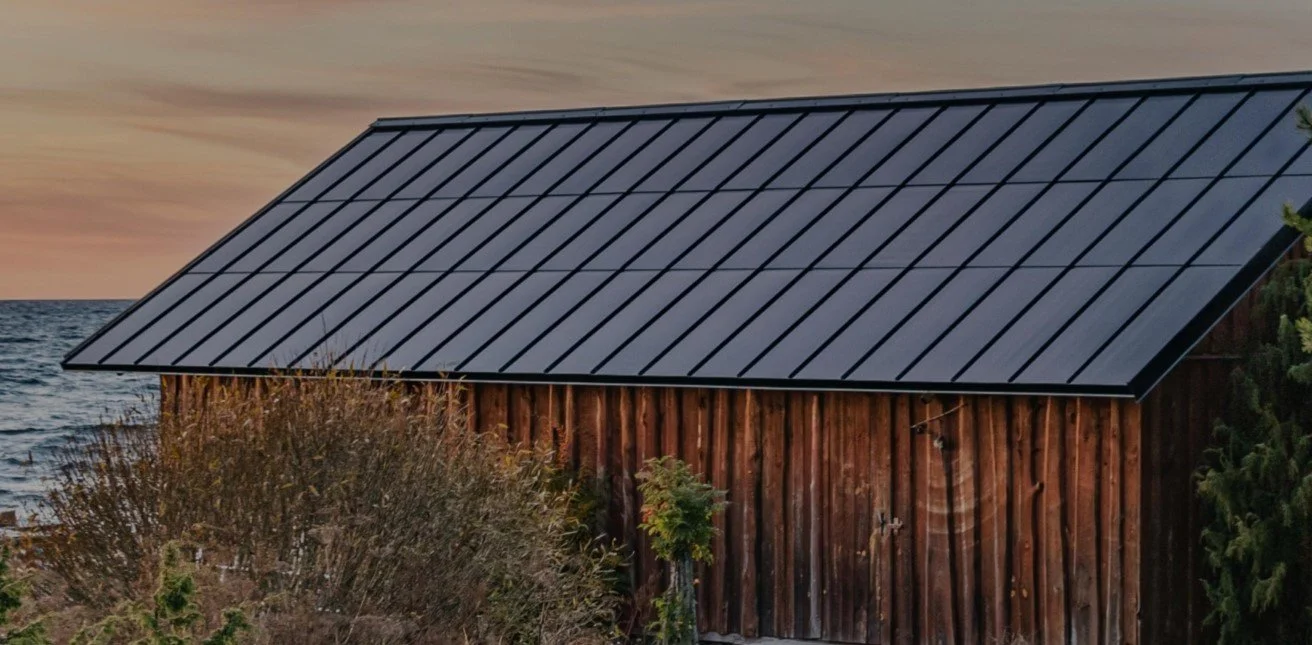 A rustic wooden building with a black metal roof beside a body of water, seen at sunset with a colorful sky.
