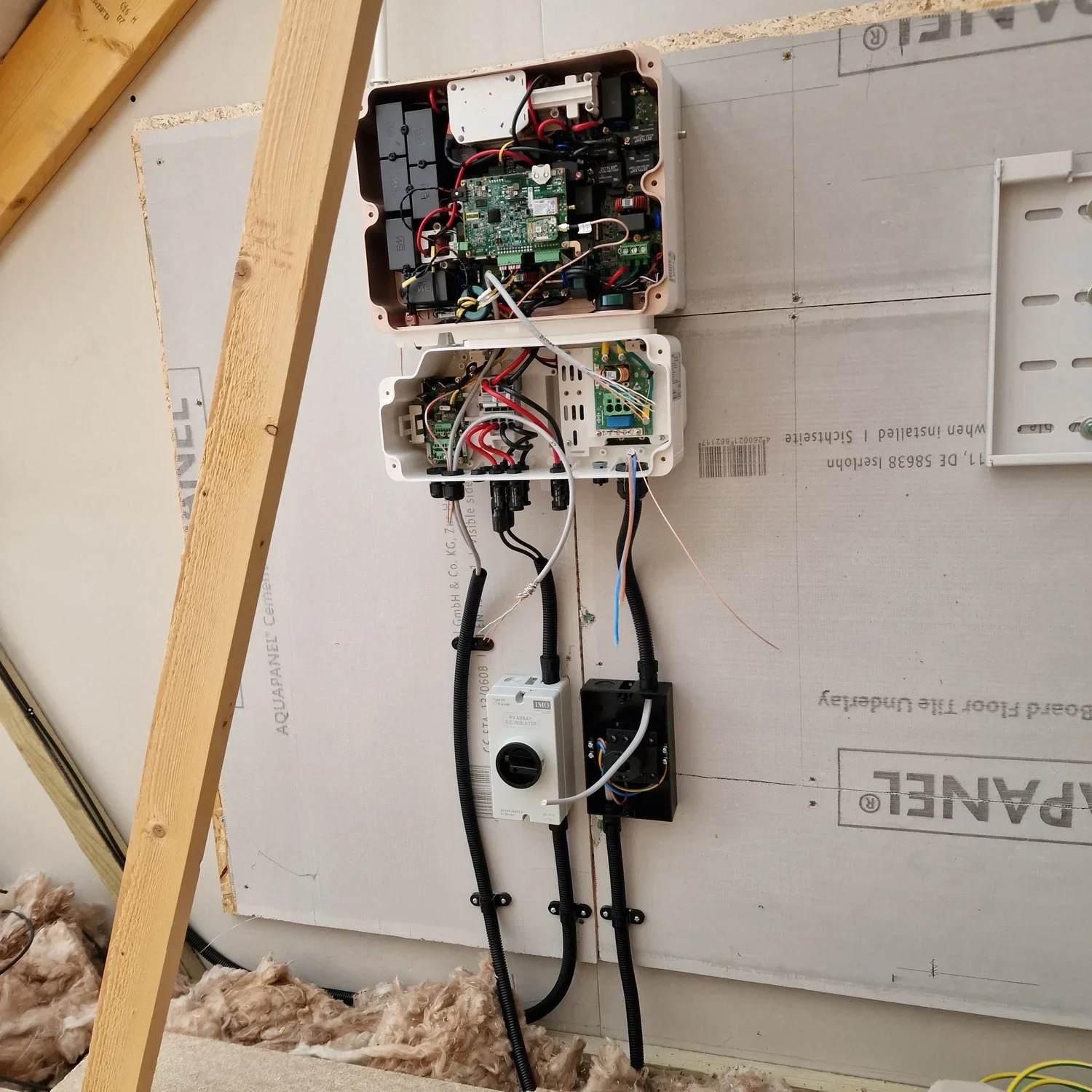 Electrical control boxes mounted on a wall, with wires and components inside. One is open, revealing circuit boards and wiring, with cables running down to a switch or breaker box.