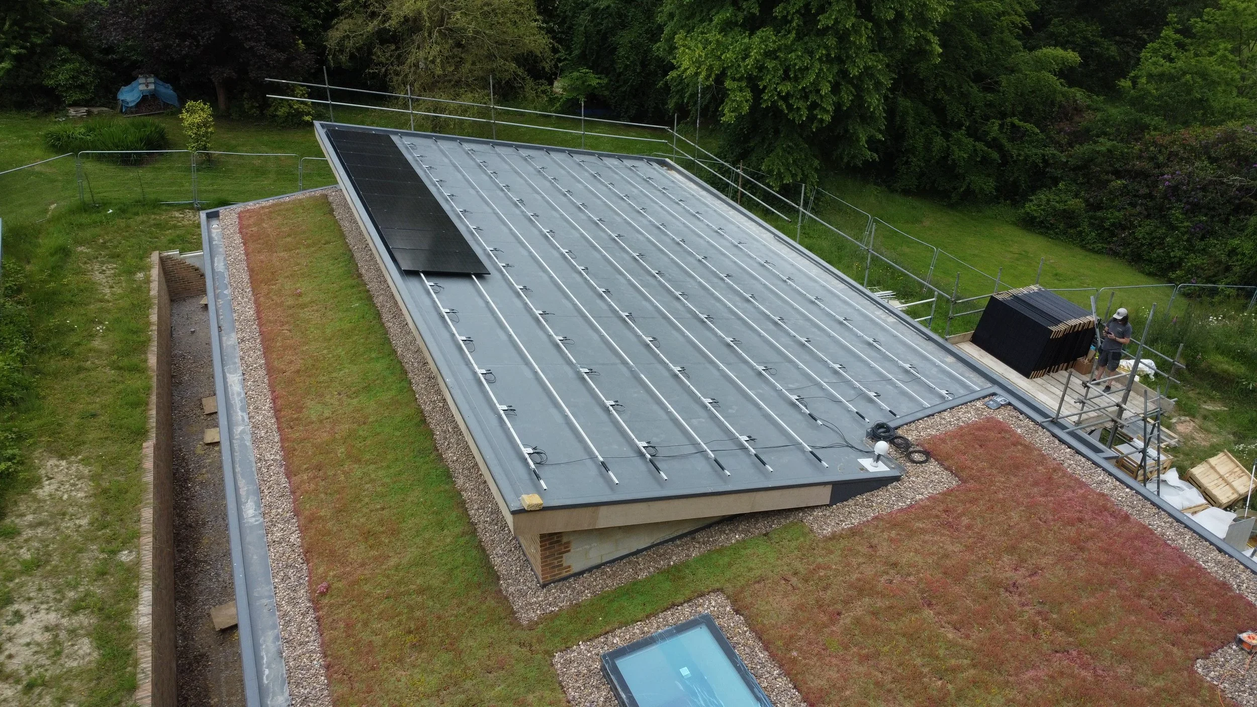 A rooftop with solar panels being installed, surrounded by green trees and a grassy yard, with construction workers working on the roof.