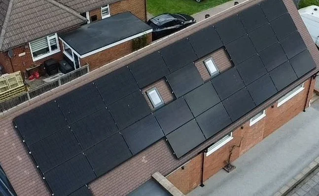 Aerial view of a residential rooftop covered with black solar panels, two skylights, and neighboring houses and cars visible in the background.