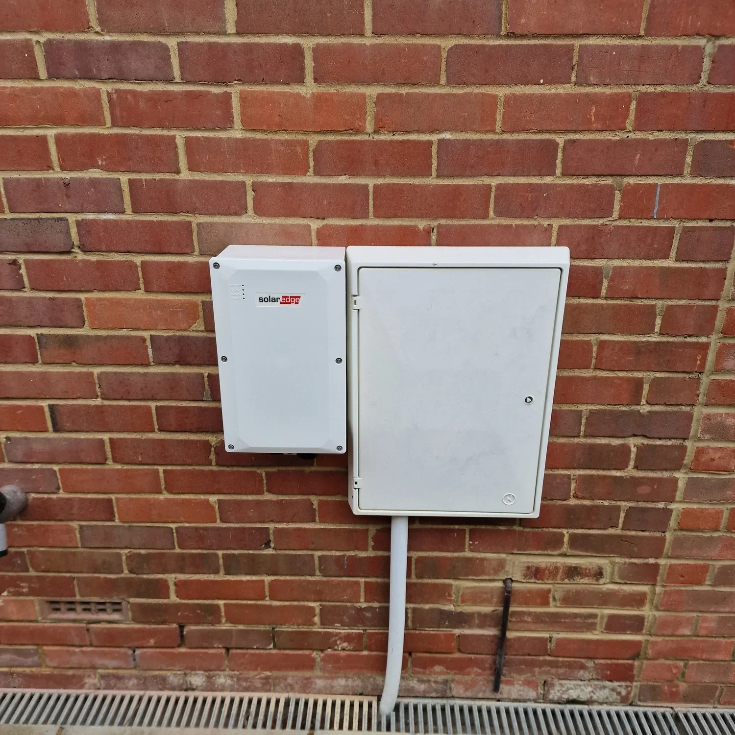 A brick wall with two electrical boxes, one labeled "solaredge," mounted on it. A conduit runs from the bottom of the smaller box downwards, and a black rod is at the base of the wall.