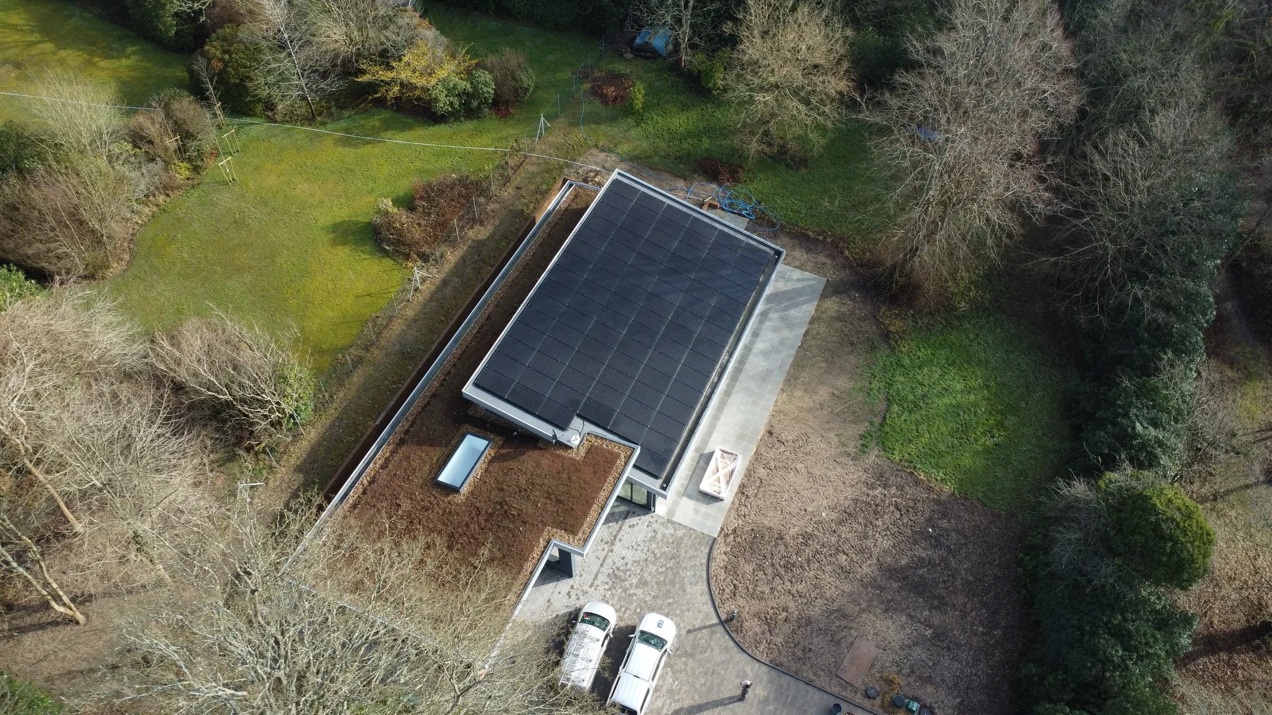 Aerial view of a house with solar panels on the roof, surrounded by trees, a driveway with two cars parked, and a backyard with grass and trees.