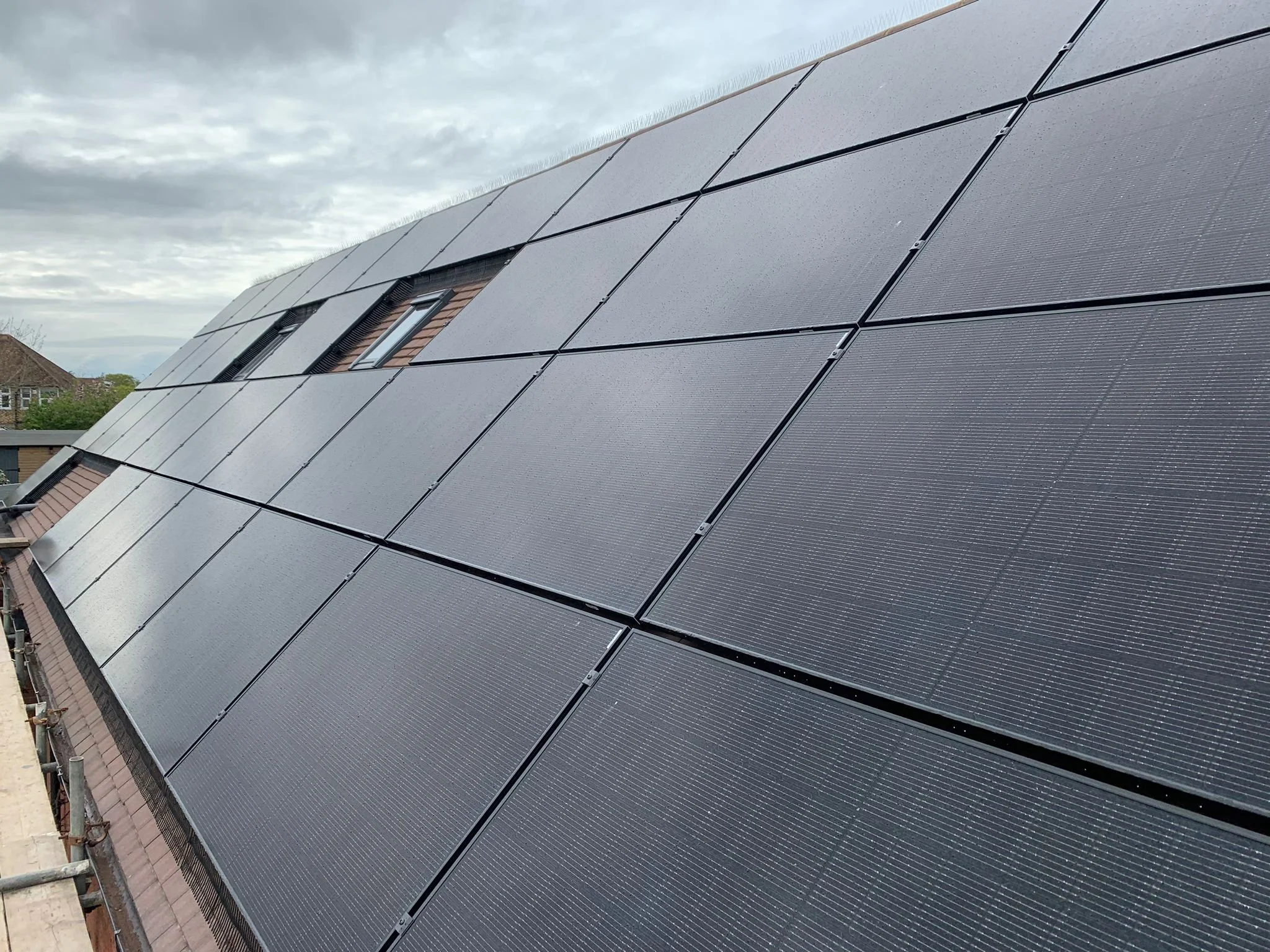A rooftop covered with black solar panels installed over a sloped roof, with a small window visible among the panels, under a cloudy sky.