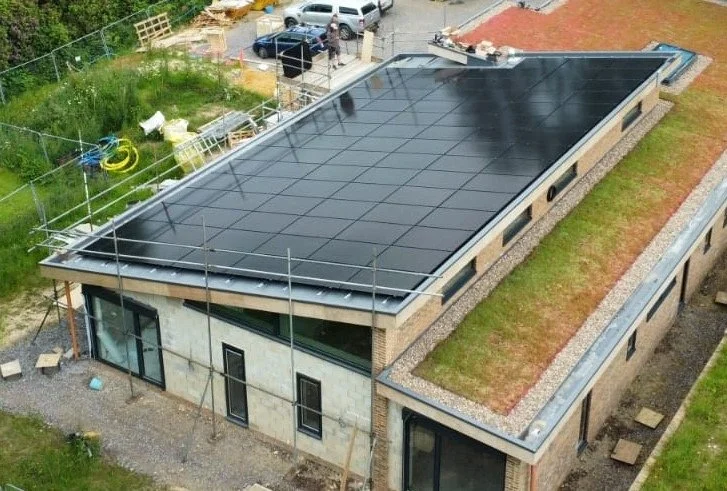 A modern two-story house under construction with a large flat roof covered in black solar panels, surrounded by construction materials and a parking area.