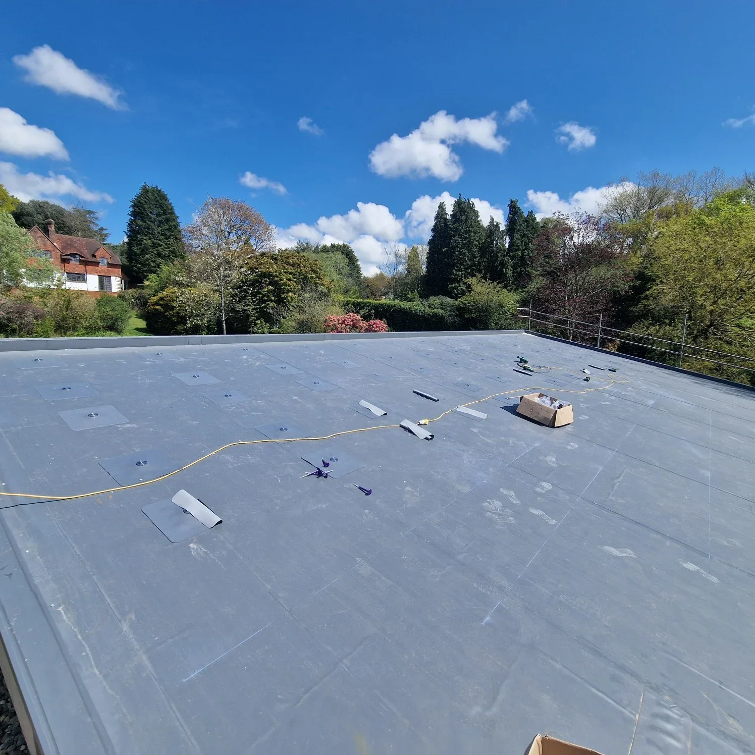 Photo of a flat roof with tools and materials for roofing work, set against a backdrop of trees and houses under a blue sky with scattered clouds.